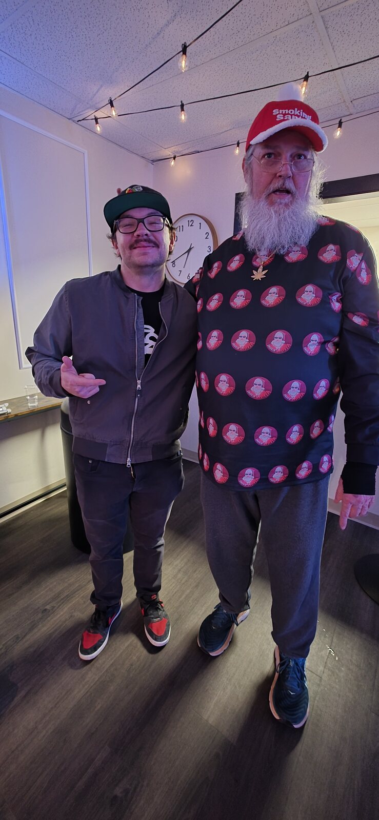 Smoking Santa in a shirt printed with his own face, beside a shop worker under festoon lights