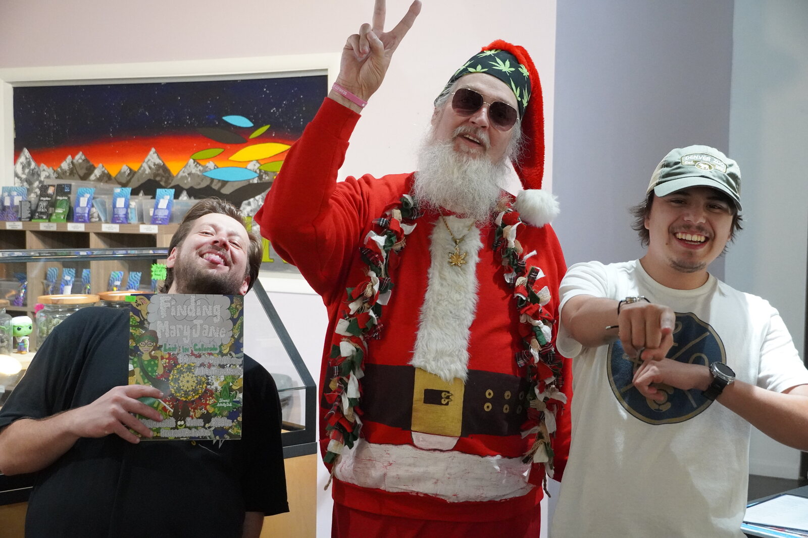 Smoking Santa flashing a peace sign with two dispensary staff holding a Finding Mary Jane activity book