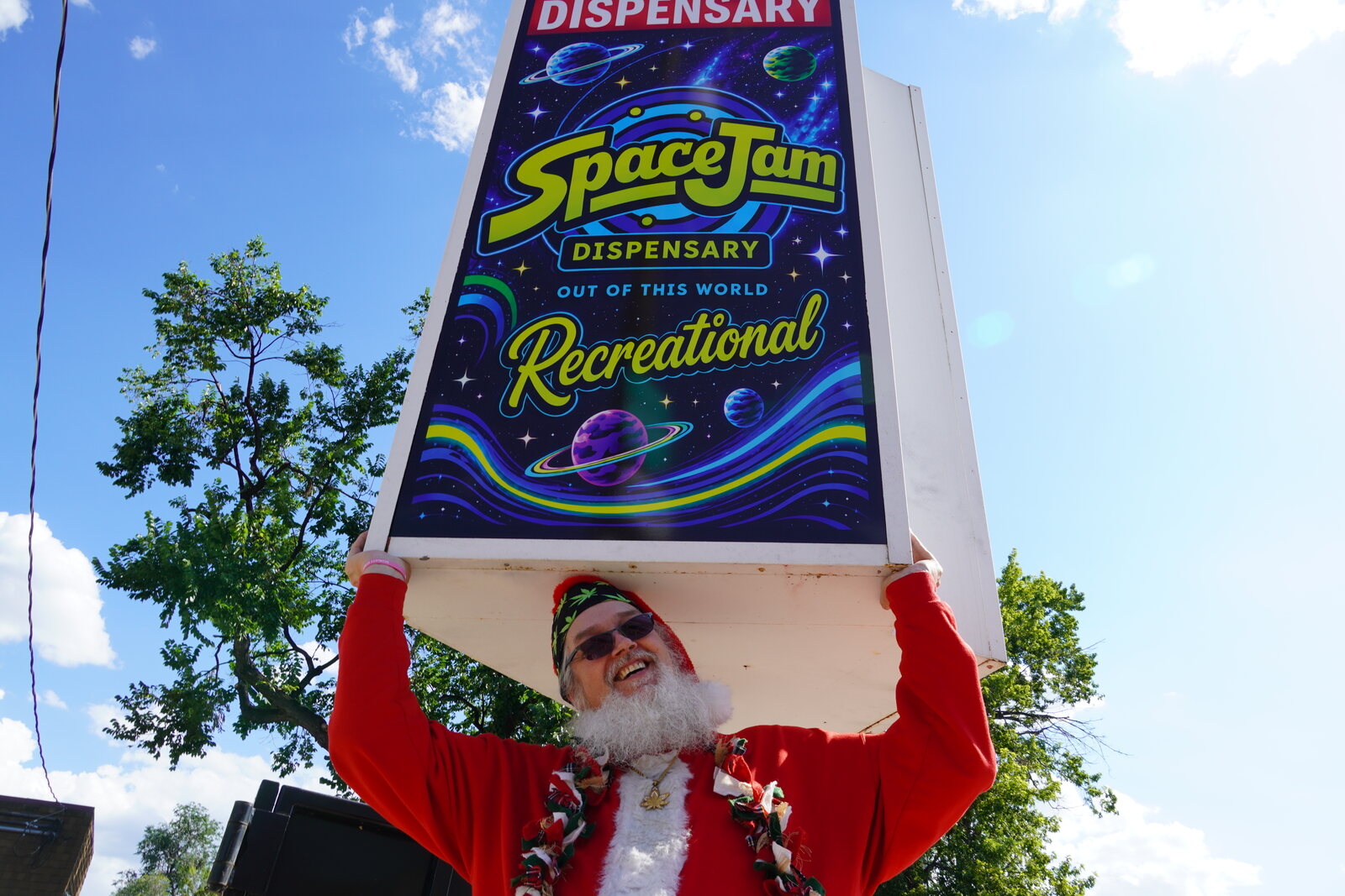 Smoking Santa holding up the Space Jam Dispensary road sign on a summer afternoon